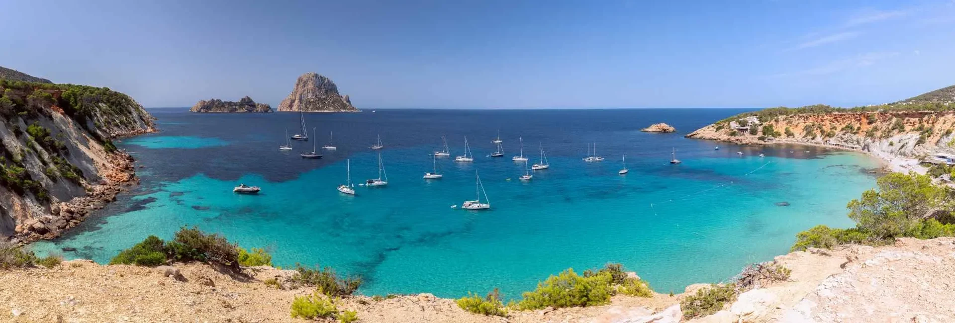 Panoramic view of the Mediterranean Sea from Ibiza’s rocky coastline with luxury yachts anchored in the bay and the iconic Es Vedrà rock formation on the horizon.