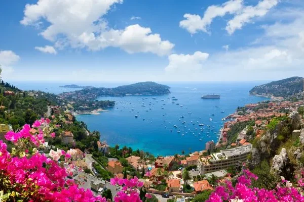 Panoramic view of the French Riviera coastline with luxury yachts anchored in the bay of Villefranche-sur-Mer and colorful Mediterranean village facades.