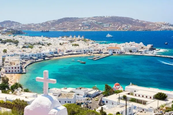 Mythical view of Mykonos windmills overlooking Little Venice, with whitewashed houses and the deep blue Aegean Sea under a clear sky.